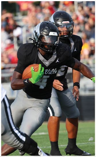 Two people in football uniforms are actively playing on a field, with one holding a football and wearing bright green gloves. Descripci n generada con IA