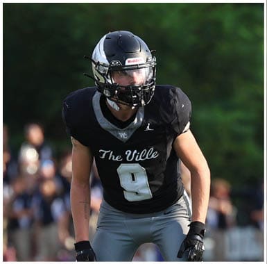 A person wearing a black football uniform and helmet stands on a green field with a crowd and trees in the background. Descripci n generada con IA