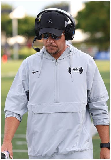 A person wearing a light gray Nike jacket with a headset, sunglasses, and a black cap stands outdoors on a sports field. Descripci n generada con IA