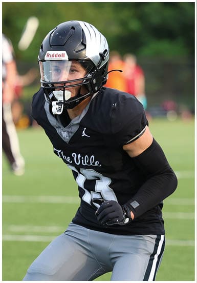 A person wearing a black football helmet and black jersey with the text “The Ville" is focused and ready to play on a green field. Descripci n generada con IA