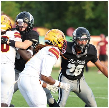 Two people in football uniforms and helmets are engaged in a play on a grassy field, with one wearing a black jersey labeled “The Ville" and the other in a white jersey with a yellow helmet. Descripci n generada con IA