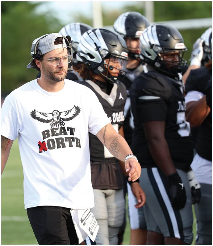A person wearing a white t-shirt with a graphic and text that says “BEAT NORTH" and a backward cap is standing on a sports field next to another person in football gear. Descripci n generada con IA