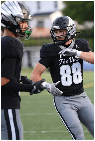 A person wearing a black football jersey with the number 88 and gray pants is preparing to throw a football on a sports field. Descripci n generada con IA