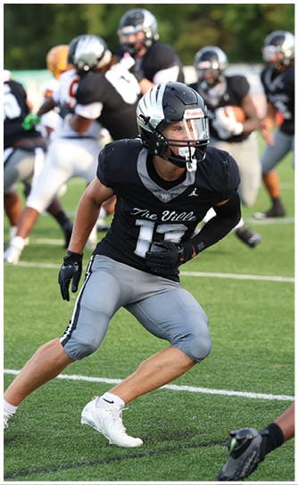 A person in a black and gray football uniform with the number 15 is crouched and ready to move on a green field with other players in the background. Descripci n generada con IA