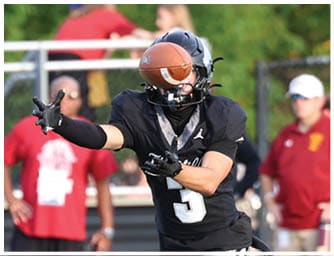 A person in a black sports uniform is throwing a football on a green field while another person in a similar uniform prepares to catch it, with several people and cheerleaders standing in the background. Descripci n generada con IA