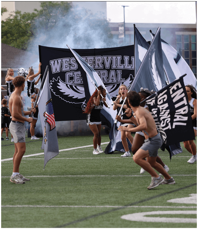 People on a sports field hold flags and a large banner that reads “Westerville Central" with cheerleaders and smoke in the background. Descripci n generada con IA