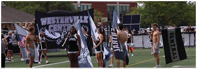 A group of people holding flags and a large banner that reads “Westerville Central" are gathered outdoors near a fence with trees and buildings in the background. Descripci n generada con IA