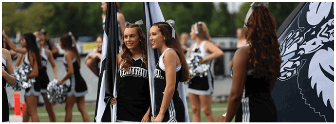 People in cheerleading uniforms are standing on a field holding flags and pom-poms, with some smiling and interacting. Descripci n generada con IA