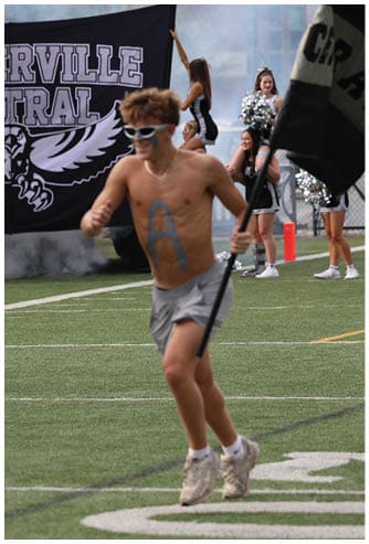 A shirtless person with painted body running on a sports field holding a flag while others in cheerleading uniforms wave flags and stand in the background near a banner that reads Westerville Central. Descripci n generada con IA