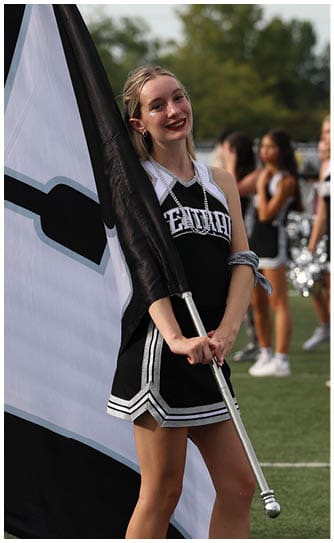 A person in a black and white cheerleading uniform is smiling and holding a large black and white flag on a sports field with other cheerleaders in the background. Descripci n generada con IA