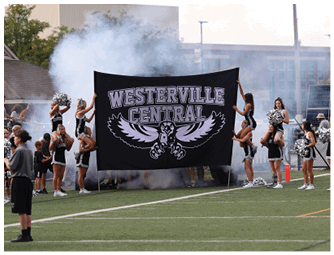 People in cheerleading uniforms hold a large banner that reads “Westerville Central" on a sports field with smoke in the background. Descripci n generada con IA