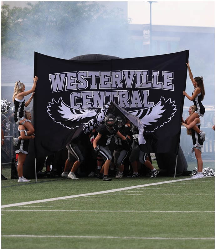 People in football uniforms are running through a large banner that reads “Westerville Central" while cheerleaders hold the banner on a sports field. Descripci n generada con IA