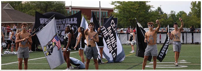 Adults wearing shorts and sunglasses are standing on a sports field holding flags with text and symbols while people and cheerleaders are visible in the background. Descripci n generada con IA