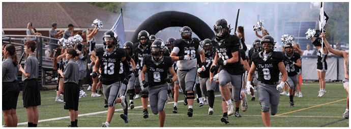 A group of people in black and gray football uniforms are running onto a field while cheerleaders in gray outfits with pom-poms stand on either side. Descripci n generada con IA
