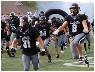 People wearing black and gray football uniforms with “The Ville" on their jerseys are running on a football field during what appears to be a team entrance or warm-up. Descripci n generada con IA