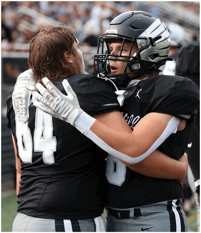 Two people in black football uniforms and helmets embrace on the field while other players stand nearby. Descripci n generada con IA