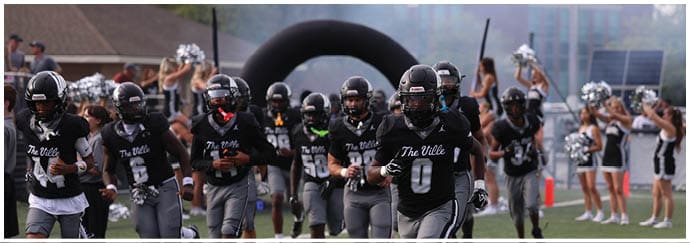 A group of people in black and gray football uniforms are running onto a field while cheerleaders in black and white outfits cheer in the background. Descripci n generada con IA