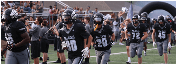 People wearing black football uniforms with helmets are running onto a field with a crowd of spectators in the background. Descripci n generada con IA