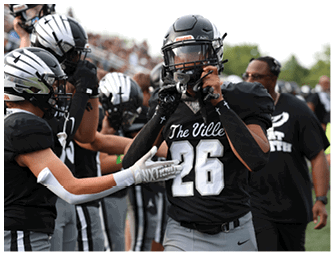 A group of people wearing black and gray football uniforms are on a field, with one person in the center adjusting their helmet while others reach out to touch or support them. Descripci n generada con IA