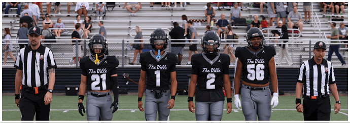 Five people wearing football uniforms and two referees in striped shirts stand on a football field with bleachers and spectators in the background. Descripci n generada con IA
