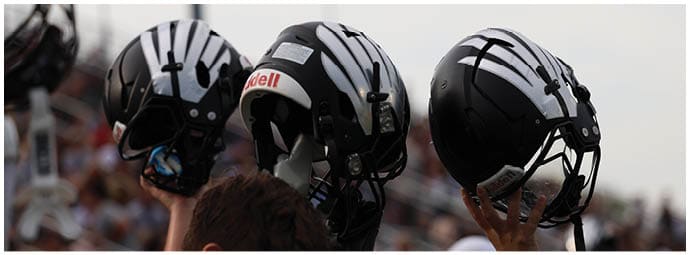 People in a stadium hold up black football helmets with white stripes against a cloudy sky. Descripci n generada con IA