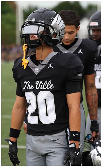 A person wearing a black football uniform with the number 20 and the words The Ville stands on a field with two other similarly dressed people in the background. Descripci n generada con IA