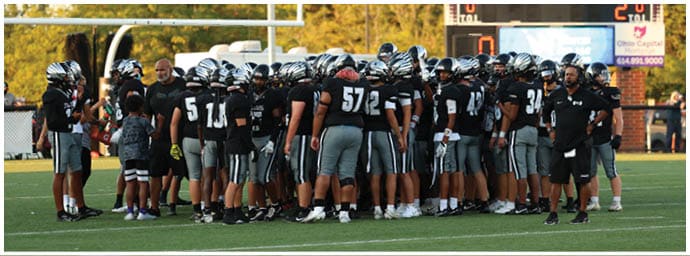 A group of people wearing black and white football uniforms and helmets are gathered on a field near a scoreboard and goalpost. Descripci n generada con IA