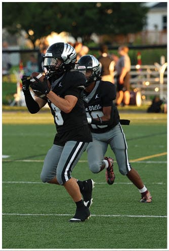 Two people in black and gray football uniforms are on a green field, with one catching a football and the other running behind. Descripci n generada con IA