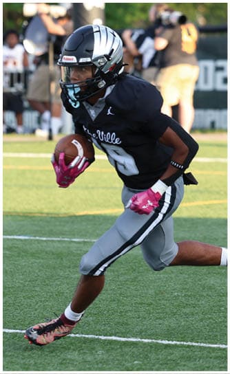 A person wearing a black football uniform and helmet is running on a green field while holding a football. Descripci n generada con IA
