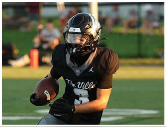 A person wearing a black football uniform and helmet is running on a green field while holding a football. Descripci n generada con IA
