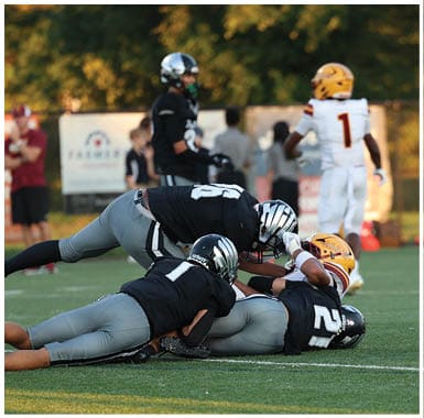 Two people in black and gray football uniforms are tackling a person in a yellow helmet and white uniform on a football field while other people stand in the background. Descripci n generada con IA