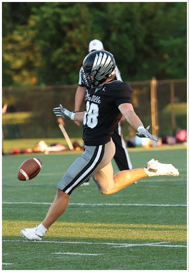 A person in a black football uniform with silver stripes is kicking a football on a green field with a blurred background of trees and a fence. Descripci n generada con IA