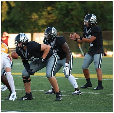 People wearing football helmets and uniforms are positioned on a field, preparing for a play during a game. Descripci n generada con IA
