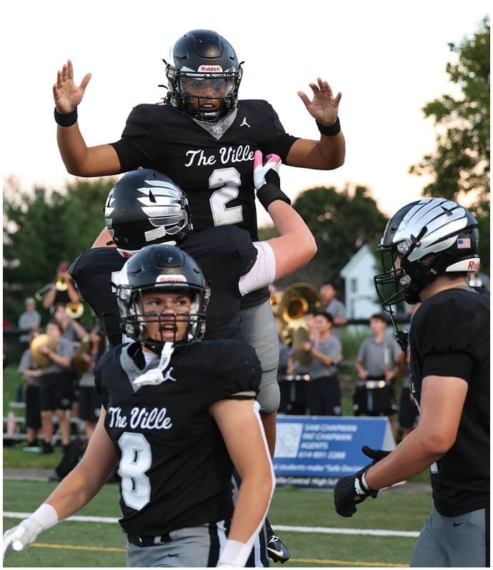 People in black football uniforms celebrating on a field with a referee nearby and a band playing in the background. Descripci n generada con IA