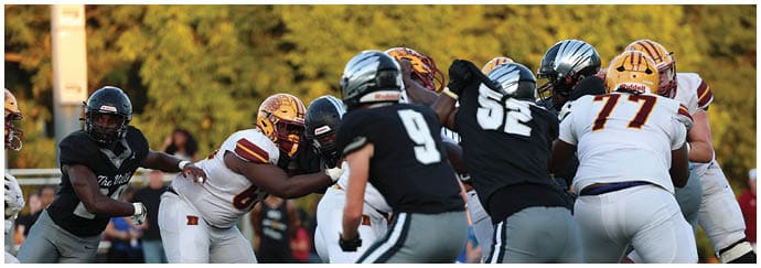 People wearing football helmets and uniforms are engaged in a play on a grassy field with trees in the background. Descripci n generada con IA