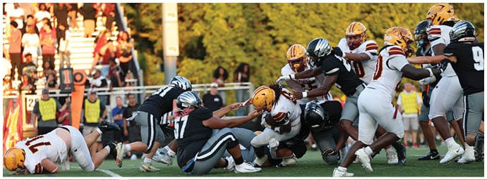 People in football uniforms are engaged in a tackle on a grassy field during a game with spectators in the background. Descripci n generada con IA