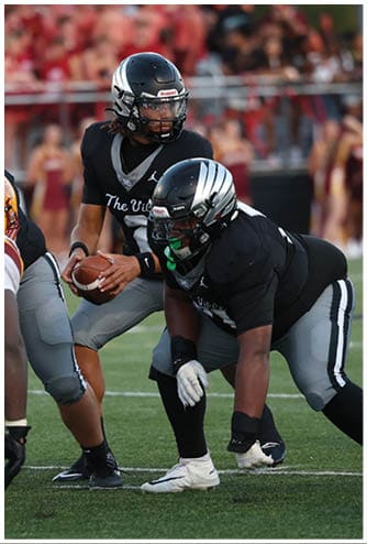 Two people in black and gray football uniforms with helmets are positioned on a football field, with one holding the ball and the other crouched in front, while an opposing player in a white and yellow uniform approaches. Descripci n generada con IA