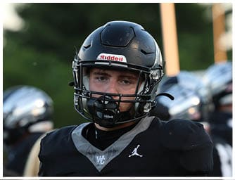 A person wearing a black football helmet and uniform is looking ahead with a blurred background showing another person in a silver helmet. Descripci n generada con IA