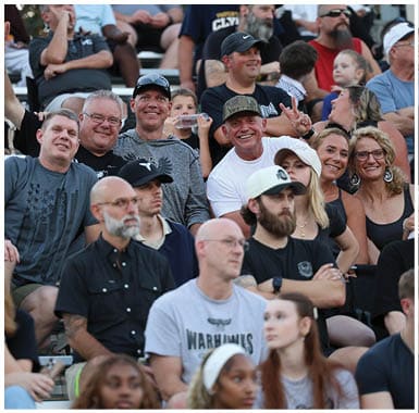 A group of adults sitting closely together on bleachers with one person making a peace sign and others smiling, while a child drinks from a cup in the background. Descripci n generada con IA