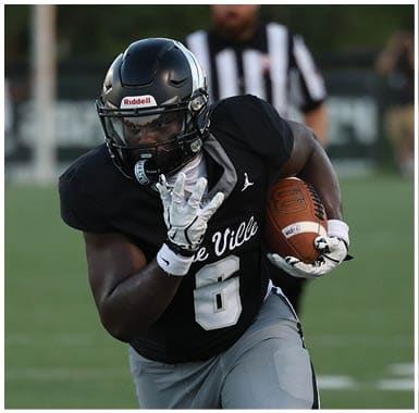 A person wearing a black football uniform with the number 6 is running on a grassy field while holding a football. Descripci n generada con IA