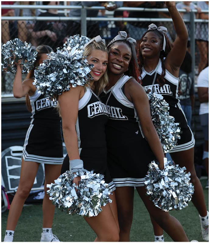 A group of smiling people in cheerleading uniforms hold silver pom-poms and pose together at an outdoor event with a crowd in the background. Descripci n generada con IA