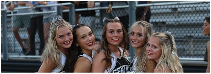 Five adults wearing cheerleading uniforms and holding silver pom-poms pose closely together outdoors near bleachers while another adult stands nearby in a skirt and top. Descripci n generada con IA