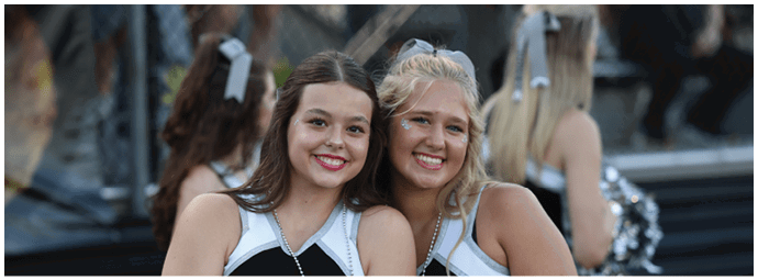 Two adults wearing cheerleading uniforms smile closely together outdoors with other cheerleaders in the background. Descripci n generada con IA