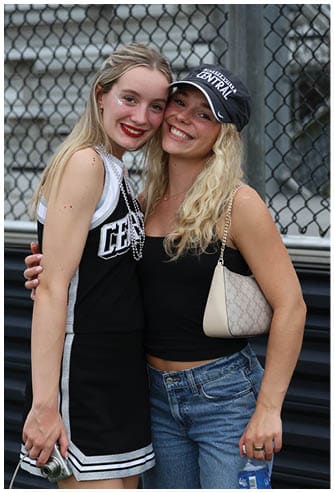 A person with long wavy hair wearing a black tank top, denim shorts, a black cap with white text, and carrying a beige shoulder bag is smiling in front of a chain-link fence. Descripci n generada con IA