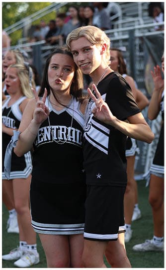 Two young people wearing cheerleading uniforms smile and pose with peace signs in an outdoor setting with a blurred crowd in the background. Descripci n generada con IA