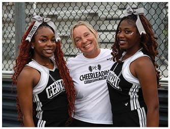 Three people smiling and posing together in cheerleading uniforms and a cheerleading-themed shirt in front of a chain-link fence. Descripci n generada con IA