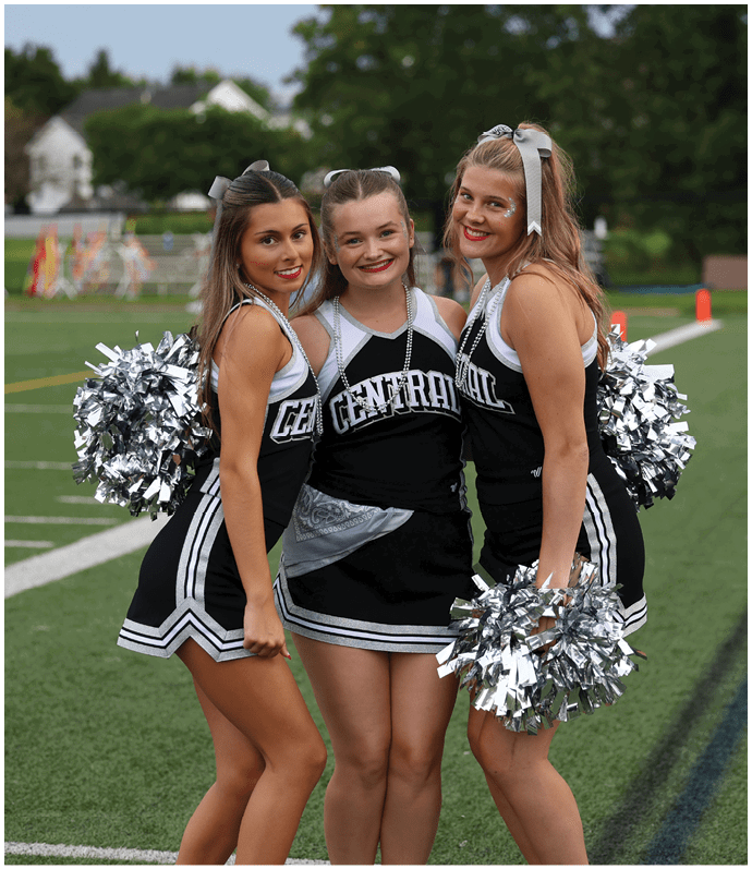 Three adults in cheerleading uniforms with silver pom-poms smile and pose closely together on a grassy field. Descripci n generada con IA