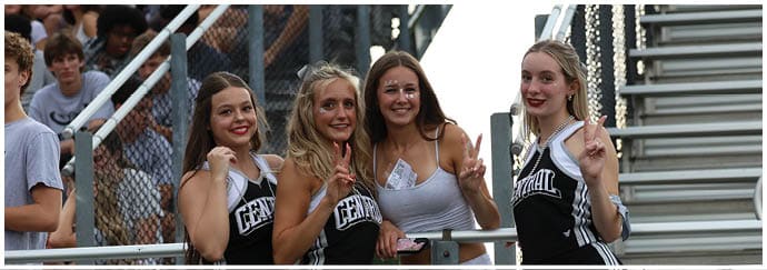 A group of adults wearing black and white cheerleading uniforms pose and smile near a chain-link fence with bleachers in the background. Descripci n generada con IA