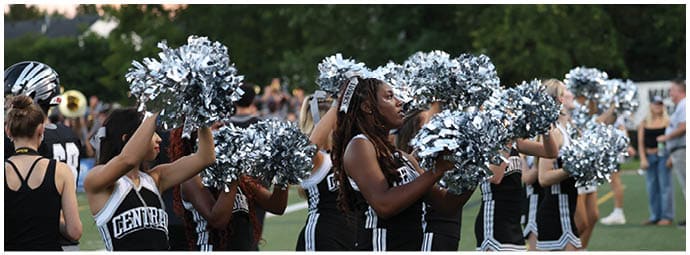 A group of cheerleaders in black and white uniforms holding silver pom-poms are performing on a grassy field with people watching in the background. Descripci n generada con IA