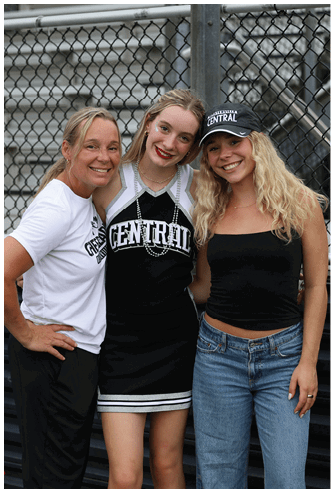 Three smiling people stand closely together in front of a chain-link fence, with one wearing a cheerleading uniform and another wearing a black tank top and jeans with a hat that says “Central." Descripci n generada con IA